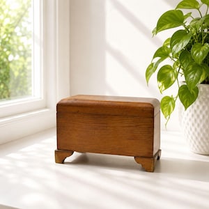 Peut inclure: Une boîte rectangulaire en bois avec une finition brun moyen lisse et un couvercle à charnière. La boîte présente un bord supérieur incurvé et repose sur quatre petits pieds sculptés à chaque coin, révélant un grain de bois naturel.