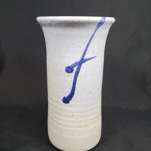May include: A tall, off-white ceramic vase with a speckled texture and a flared top. A bold, blue abstract design sweeps down the side. The vase is approximately 8 inches tall.