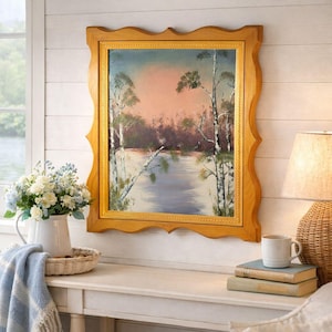 May include: A framed landscape painting with a serene lake scene, featuring birch trees and a soft, colorful sky. The painting is in a decorative, light-brown wooden frame. A white pitcher of flowers and a lamp are nearby.