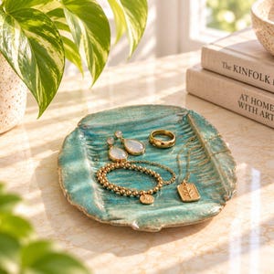 Peut inclure: Un vide-poche en céramique turquoise de forme ovale avec une surface texturée et nervurée. Sur le plat reposent une paire de boucles d'oreilles en forme de goutte, une bague anneau en or, un bracelet de perles dorées et un collier pendentif.
