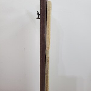 Puede incluir: Vista vertical de un marco de madera, con un borde marr&oacute;n oscuro y un panel de madera claro, posiblemente sin terminar. Una peque&ntilde;a bisagra met&aacute;lica oscura es visible en la parte superior. El marco est&aacute; contra un fondo blanco liso.