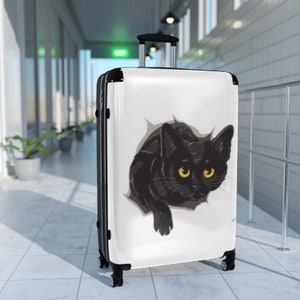 May include: White suitcase with a black cat peeking out of a hole. The cat has yellow eyes and is looking directly at the camera.
