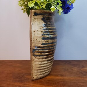 K&ouml;nnte beinhalten: Eine handgefertigte Keramikvase in Beige- und Braunt&ouml;nen. Die Vase hat eine strukturierte Oberfl&auml;che mit horizontalen Rillen und dunkelblauen Akzenten. Sie enth&auml;lt gr&uuml;ne und blaue Blumen, die einen lebendigen Kontrast bilden. Ein dekorativer Gegenstand.