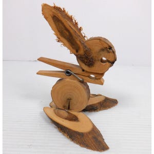 Puede incluir: Una escultura de madera caprichosa de un pájaro, hecha de rodajas de madera natural y una pinza de ropa. El pájaro presenta una cabeza circular, una pieza en forma de ala y un cuerpo sostenido por una pinza de ropa, con dos pies de madera. Una pieza única de decoración del hogar.