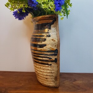 K&ouml;nnte beinhalten: Eine handgefertigte Wandvase aus Keramik mit einem einzigartigen, strukturierten Design. Die Vase weist horizontale B&auml;nder aus schwarzer und brauner Glasur auf einem beigen Grund auf. Sie enth&auml;lt eine lebendige Anordnung aus blauen und gr&uuml;nen Kunstblumen und verleiht jedem Raum Farbe.