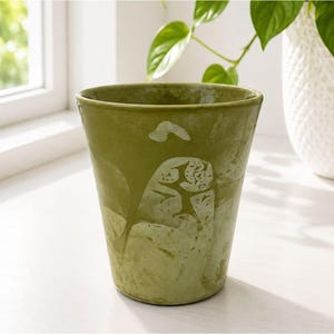 May include: A tapered ceramic planter in a muted olive green hue with a smooth, glossy finish. The surface features a subtle, abstract marbled pattern in lighter shades of green, creating a soft, organic visual texture across the exterior.