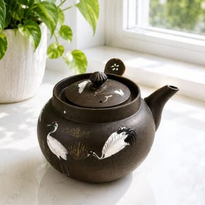 Puede incluir: Una tetera de cerámica marrón oscuro con tapa y boquilla. La tetera está decorada con ilustraciones de grullas blancas y un diseño floral en la tapa. La tetera está sobre una superficie blanca.