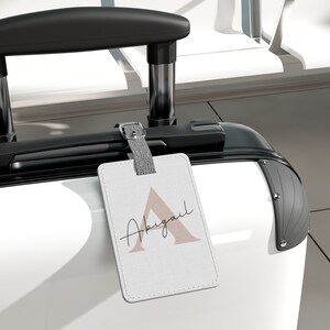 May include: White suitcase with a black handle and a luggage tag that says "Abigail" with a pink "A" in the background.