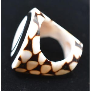 May include: A distinctive, geometric ring. It has a white base with brown, organic-shaped patterns. The ring has a bold, angular design with a smooth, polished finish. The ring's design includes a black and white striped section.