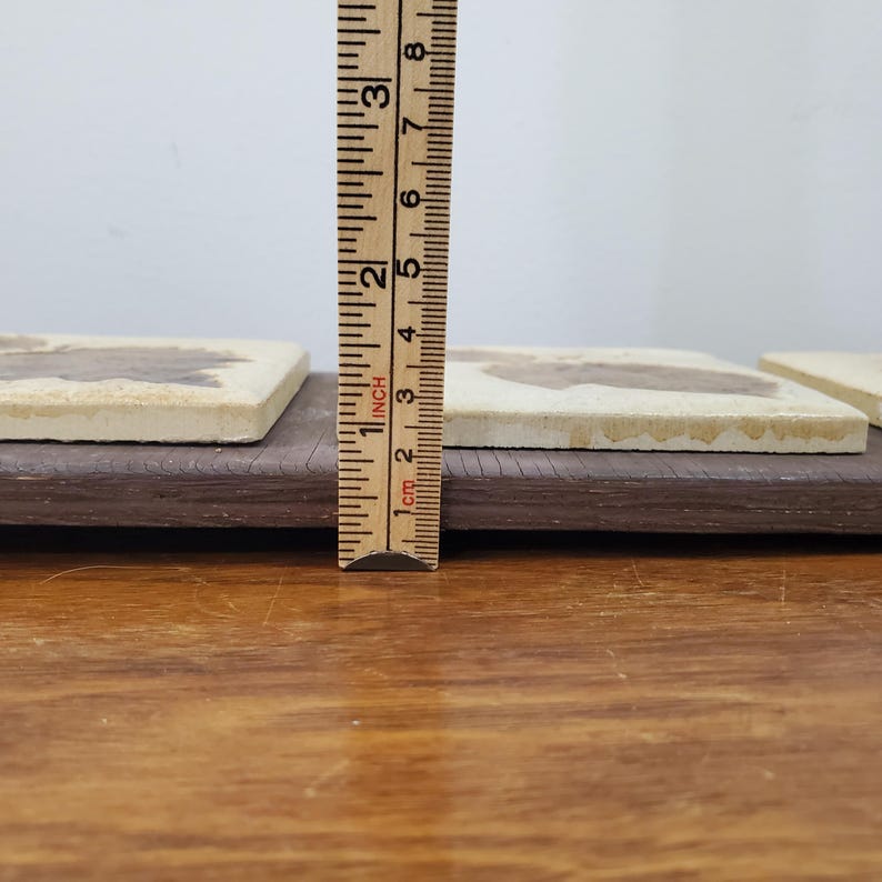 Puede incluir: Posavasos cuadrados con una superficie beige claro y un dise&ntilde;o marr&oacute;n, apoyados sobre una base de madera marr&oacute;n oscuro. Los posavasos miden aproximadamente 7,6 cm de largo, medidos con una regla.