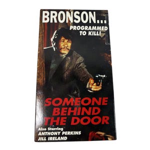 Pode incluir: Uma fita VHS vintage do filme "Someone Behind the Door" com Charles Bronson. A capa apresenta Bronson segurando uma arma, com o título do filme em texto vermelho e branco.