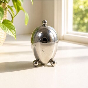 May include: A silver-colored sugar bowl with a lid and three spherical feet. The bowl has a smooth, reflective surface and a small opening for a spoon. The design is egg-shaped, with a small knob on top of the lid.