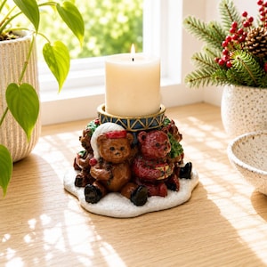 May include: A cylindrical cream-coloured pillar candle sits atop a circular base decorated with multiple brown and red teddy bears. The bears are positioned on a white textured surface resembling snow, with some wearing hats and holding small gifts.