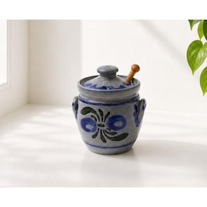 May include: A grey ceramic jar with a lid and two side handles, featuring a hand-painted blue and black floral motif. A small wooden honey dipper is inserted through a notch in the lid, resting against the rim of the textured stoneware container.