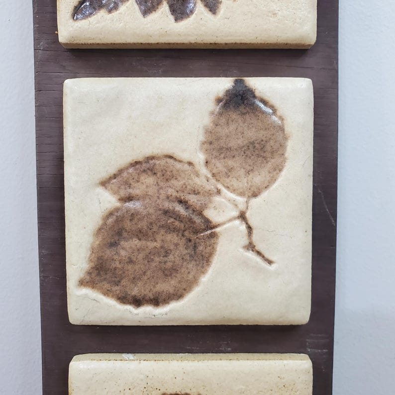 Puede incluir: Adorno de pared con baldosas de cer&aacute;mica crema con dise&ntilde;os de hojas marrones. Las hojas, con sus vetas y sombreado, est&aacute;n fijadas a un soporte de madera marr&oacute;n oscuro, creando un efecto natural.