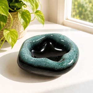 May include: A teal and black ceramic ashtray with a distinctive, organic form. The ashtray features a dark interior and a textured, teal rim. It is presented on a white surface, with a houseplant visible in the background.