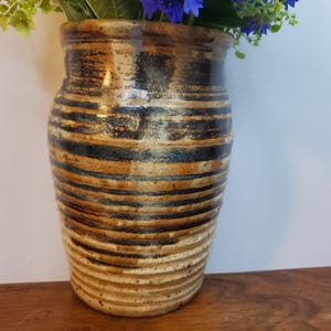 K&ouml;nnte beinhalten: Eine handgefertigte Keramikvase mit einem strukturierten, gerippten Design. Die Vase hat horizontale B&auml;nder in Braun, Schwarz und Beige. Sie enth&auml;lt blaue und gr&uuml;ne Blumen. Ein dekoratives Objekt f&uuml;r das Zuhause.