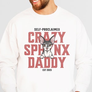May include: White sweatshirt featuring the text "SELF-PROCLAIMED CRAZY SPHINX DADDY EST 2023" in red. A cartoon Sphynx cat is in the center. The cat is grey and white with large ears. The sweatshirt is a casual style.