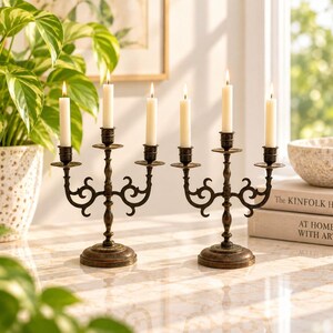 Peut inclure: Une paire de candélabres en métal de couleur bronze foncé, chacun doté de trois bras avec des détails en volutes incurvées et des bases circulaires. Chaque candélabre porte trois bougies effilées blanches, formant un ensemble de six bougies.