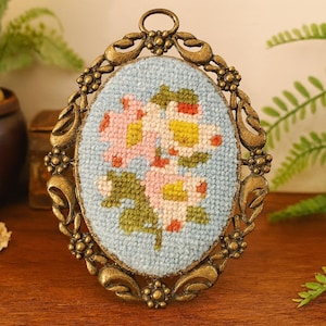 Könnte beinhalten: Ein ovaler, vintage-Stil gerahmter Nadelpunkt mit einem Blumendesign. Der Nadelpunkt hat einen hellblauen Hintergrund mit rosa, gelben und grünen Blumen. Der verzierte Rahmen hat eine Bronzefinish und eine Schlaufe oben zum Aufhängen.
