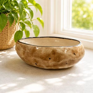 May include: A ceramic bowl with a potato-like design. The bowl is oval-shaped with a textured, brown exterior and a black rim. Small, dark spots are scattered across the surface. The interior is a light beige color.