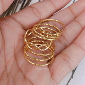 May include: A collection of gold-colored rings in various designs, including twisted and interwoven patterns. The rings are stacked together, showcasing their different textures and styles. The image is taken from a close-up angle, highlighting the details of the jewelry.