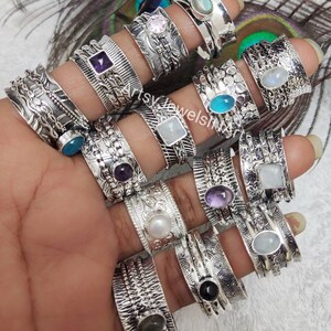 May include: A collection of 14 silver spinner rings featuring a variety of gemstones, including amethyst, moonstone, pearl, and labradorite. Each ring has intricate designs and a spinning band.