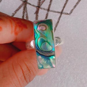 May include: A rectangular silver ring featuring an abalone shell inlay. The iridescent shell displays shades of green, blue, and purple. The ring's band is a simple, polished silver.
