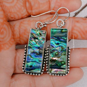 May include: Silver-tone rectangular earrings with iridescent abalone shell in shades of blue, green, and purple. The earrings have a beaded border and a hook closure. The text "ArtsyJewelsINArt" is visible.