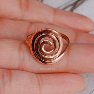 May include: A copper-colored ring with a spiral design. The ring is held in a hand, showcasing the intricate swirl pattern. The ring's design is centered, with the band curving around the finger. The ring is a unique piece of jewelry.