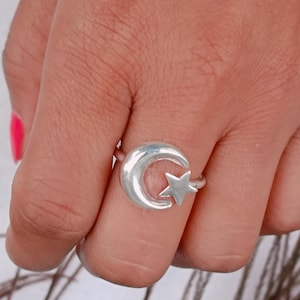 Op de afbeelding: Een zilveren ring met een halve maan en ster ontwerp. De ring is gemaakt van een glanzend, metalen materiaal. Het ontwerp van de ring heeft een hemels thema.