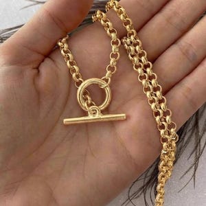 May include: A gold-toned necklace featuring a thick rolo chain and a prominent toggle clasp closure. The necklace consists of a circular ring and a horizontal bar component that interlocks to secure the chain, showcasing a polished metallic finish.