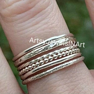 May include: A set of five silver stacking rings with different textures, including a beaded ring, a hammered ring, and a ring with a textured surface. The rings are worn on a finger.