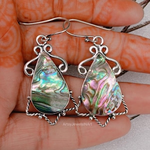 Può includere: Orecchini in argento con pendenti a goccia in conchiglia di abalone iridescente. I gusci mostrano tonalità verdi, blu, rosa e viola. Gli orecchini hanno una cornice decorativa in argento e una chiusura a gancio. Un gioiello unico.