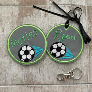 Könnte beinhalten: Zwei graue runde Anhänger mit grünem Rand, jeweils mit einem Fußball-Grafik und einem Namen in grüner Schrift: "Matteo" und "Leon". Die Anhänger haben ein schwarzes Band und einen silbernen Verschluss.
