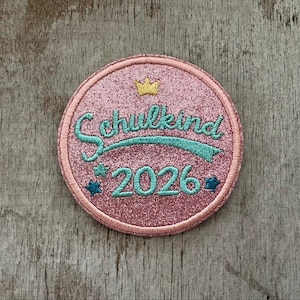 Könnte beinhalten: Ein runder, rosa Glitzeraufnäher mit dem Wort "Schulkind" in türkisfarbener Schrift, einem türkisfarbenen Banner und der Zahl "2026". Der Aufnäher hat eine goldene Krone und türkisfarbene Sternakzente. Der Aufnäher ist mit einem rosa Rand bestickt.