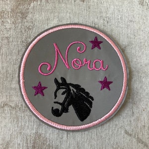 Könnte beinhalten: Ein runder grauer Patch mit rosa Rand. Der Patch zeigt eine schwarze Pferdesilhouette, den Namen "Nora" in rosa Kursivschrift und drei rosa Sterne.