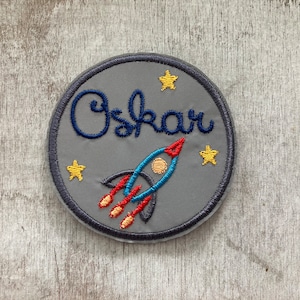 Könnte beinhalten: Ein grauer Filz-Kreis mit einem blauen gestickten Namen "Oskar" und einer roten, blauen und gelben gestickten Rakete mit drei gelben Sternen.