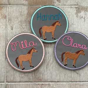 Könnte beinhalten: Drei gestickte Aufnäher mit den Namen "Hannah", "Mila" und "Clara". Jeder Aufnäher zeigt eine braune Pferdeillustration auf grauem Hintergrund, mit farbigem Rand und passendem Namen.