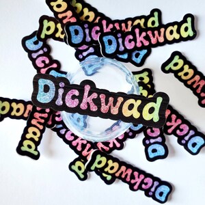 May include: A close-up of a glass jar surrounded by colorful stickers that say "Dickwad". The stickers are on a white background.