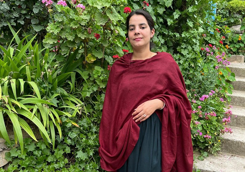 May include: A woman wearing a maroon shawl over a dark green dress. She is standing in front of a wall of green plants with pink flowers.