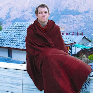 May include: A person wrapped in a dark red blanket with fringed edges, sitting outdoors. The blanket covers the person's shoulders and legs. The background features a mountain range, buildings, and trees.