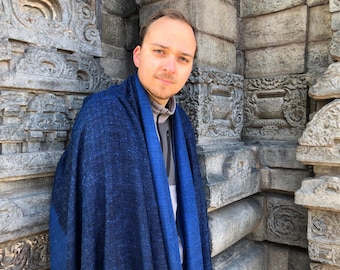 Peut inclure: Un homme portant un châle bleu et noir à motifs se tient devant un mur de pierre aux sculptures complexes. Le châle est drapé sur ses épaules et est fait d'un tissu doux et texturé.