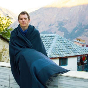 Peut inclure: Un homme portant un ch&acirc;le bleu fonc&eacute; est assis sur un rebord en marbre blanc avec une cha&icirc;ne de montagnes en arri&egrave;re-plan. L'homme regarde droit devant lui avec une expression calme.