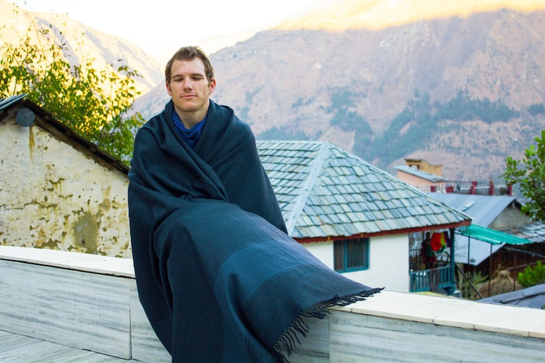 Peut inclure: Un homme portant un ch&acirc;le bleu fonc&eacute; est assis sur un rebord en marbre blanc avec une cha&icirc;ne de montagnes en arri&egrave;re-plan. L'homme regarde droit devant lui avec une expression calme.