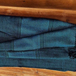 Peut inclure: Une &eacute;charpe tiss&eacute;e bleu sarcelle avec des rayures noires et des franges noires. L'&eacute;charpe est pli&eacute;e et pos&eacute;e sur une surface en bois.