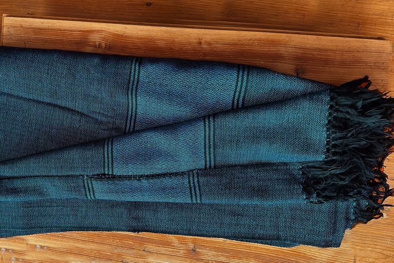 Peut inclure: Une &eacute;charpe tiss&eacute;e bleu sarcelle avec des rayures noires et des franges noires. L'&eacute;charpe est pli&eacute;e et pos&eacute;e sur une surface en bois.