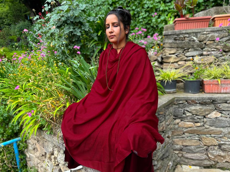 May include: A person wearing a deep red shawl and a beaded necklace, seated outdoors. The shawl drapes over the shoulders and body. The background features lush greenery, stone steps, and potted plants, creating a serene atmosphere.