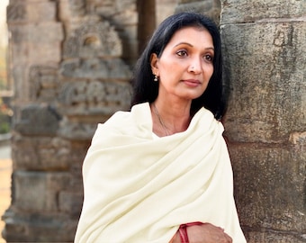 May include: A woman with dark hair and a cream-colored shawl stands near a weathered stone structure. She wears small earrings and a delicate necklace. The background features ancient stone architecture, suggesting a historical or cultural setting.