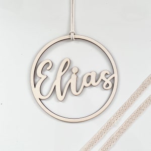 Könnte beinhalten: Ein Holz-Kreis mit dem Namen "Elias" ausgeschnitten in einer kursivschrift. Der Kreis hängt an einer weißen Schnur und befindet sich auf einem weißen Hintergrund.